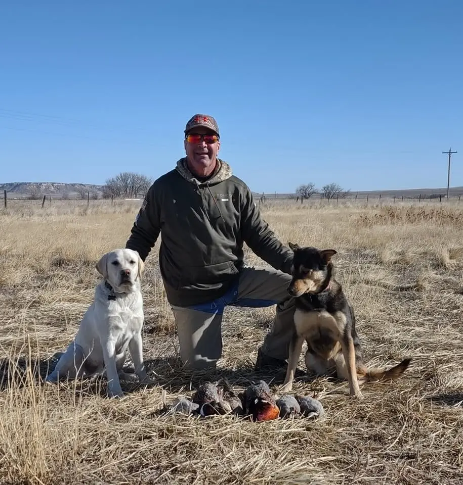 A man kneels in a dry field with two dogs and several harvested pheasants and chukar.