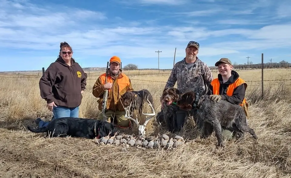 Four hunters and two dogs pose with their harvested chukar partridge in a field.