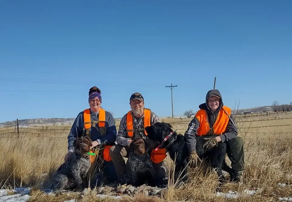 Three hunters and three hunting dogs pose in a field under a clear blue sky.
