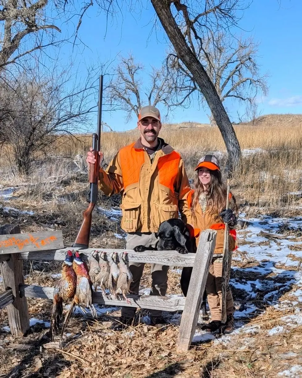 A man, a girl, and a black dog pose with harvested ring-necked pheasants and chukar partridges hanging on a fence in a snowy field.