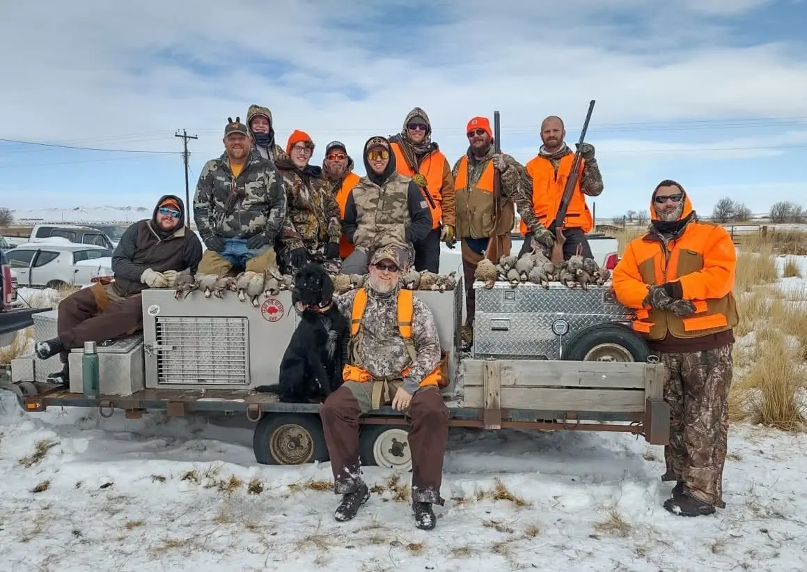 A group of hunters and dogs pose with their harvested chukar partridges on a snowy day.