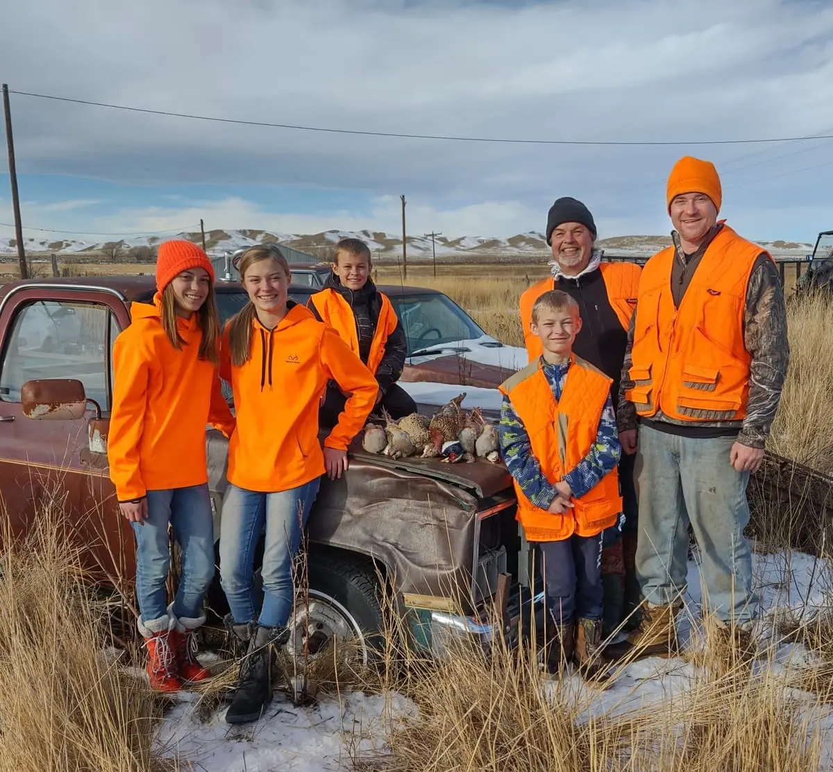 A group of hunters, including adults and children, pose with harvested pheasants and chukar next to a truck in a snowy field.