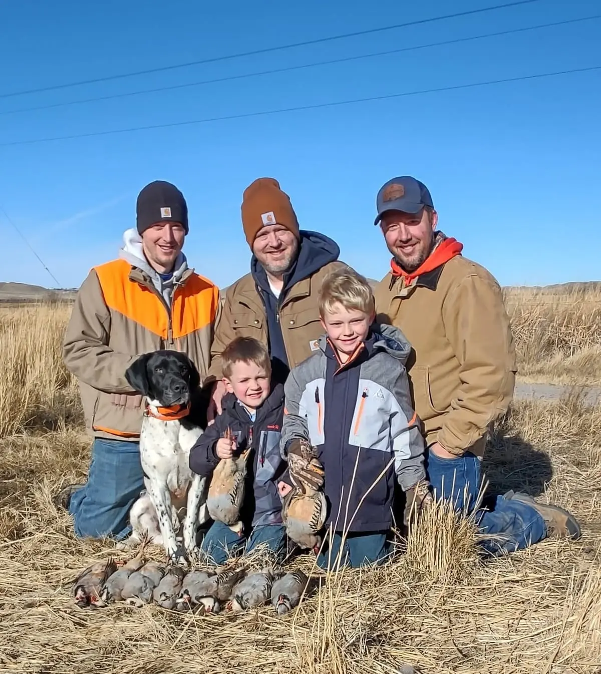 Three men, two boys, and a hunting dog pose in a field with harvested chukar partridges.