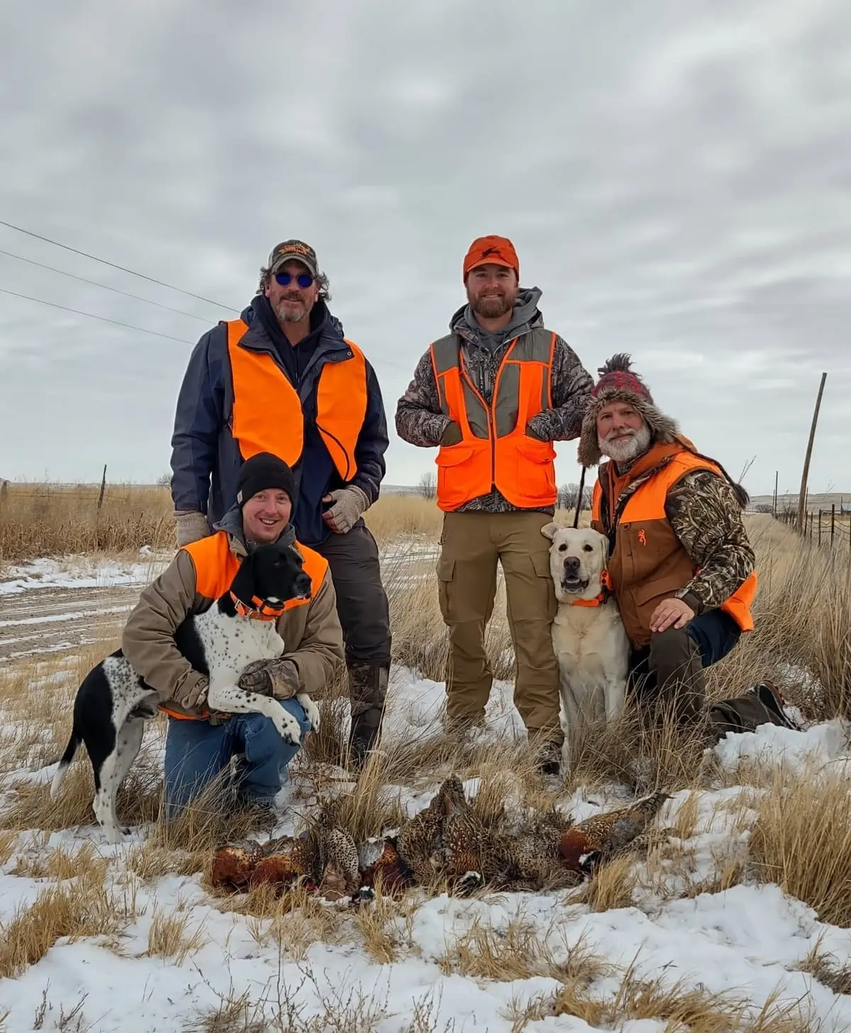 Four hunters and two dogs pose with harvested pheasants in a snowy field.