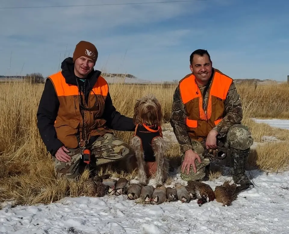 Two hunters and a dog pose with harvested Ring-necked Pheasants and Chukar Partridges in a snowy field.