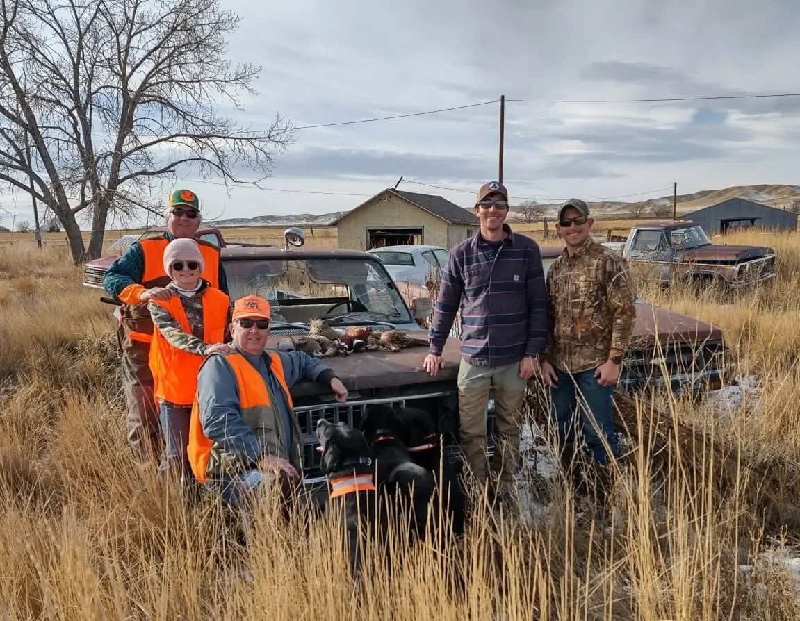 Hunters and a dog pose with harvested pheasants and chukar on the hood of a truck in a field.