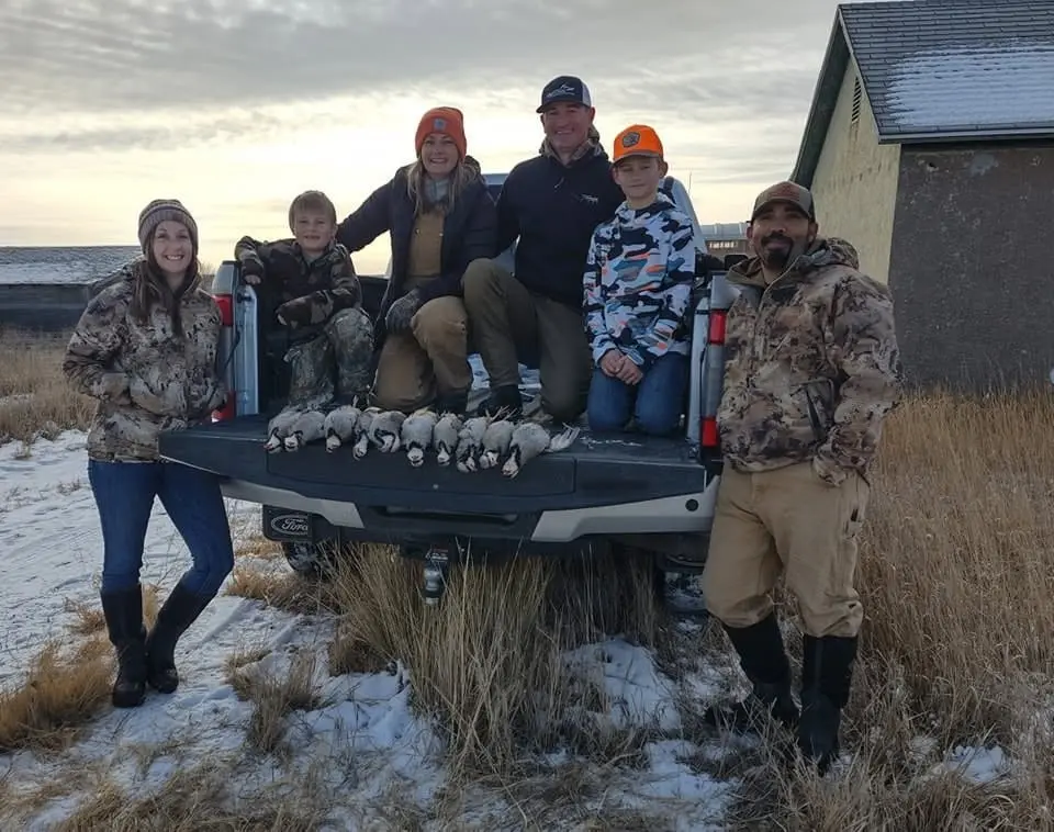 A group of hunters, including adults and children, pose with harvested birds in the back of a pickup truck in a snowy field.