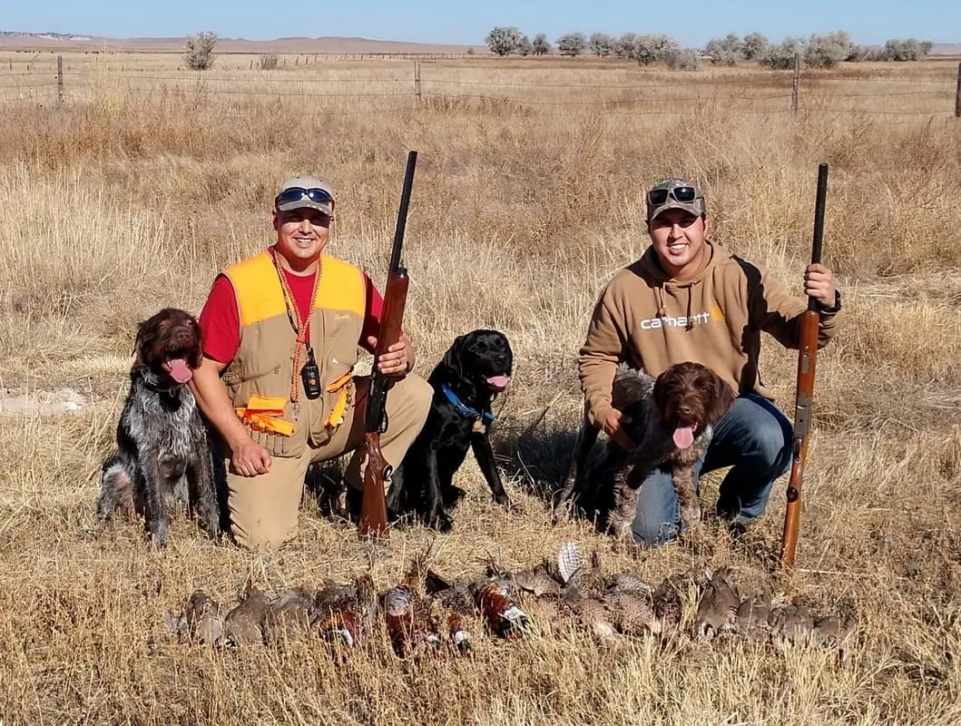 Two hunters pose with their dogs and a harvest of pheasants and chukar partridges in a field.