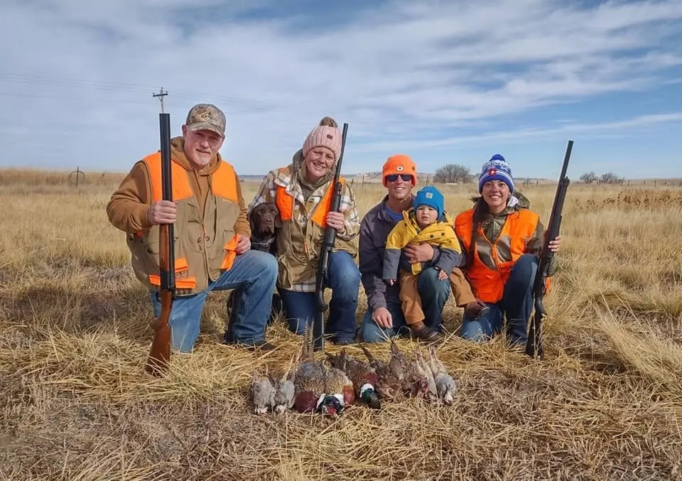 A family of hunters, including a child and a dog, pose with harvested pheasants and chukar in a field.