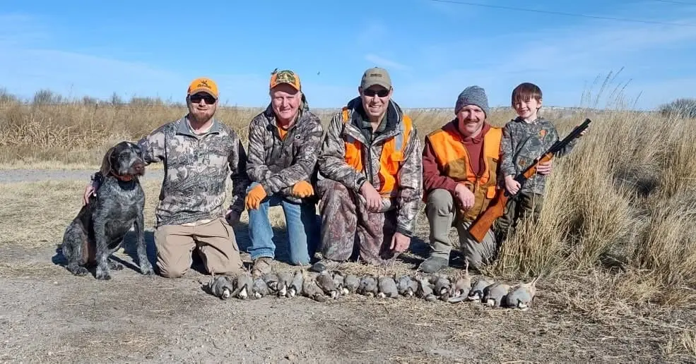 A group of hunters, including a child and a dog, pose with their harvested chukar partridges on a sunny day in a field.