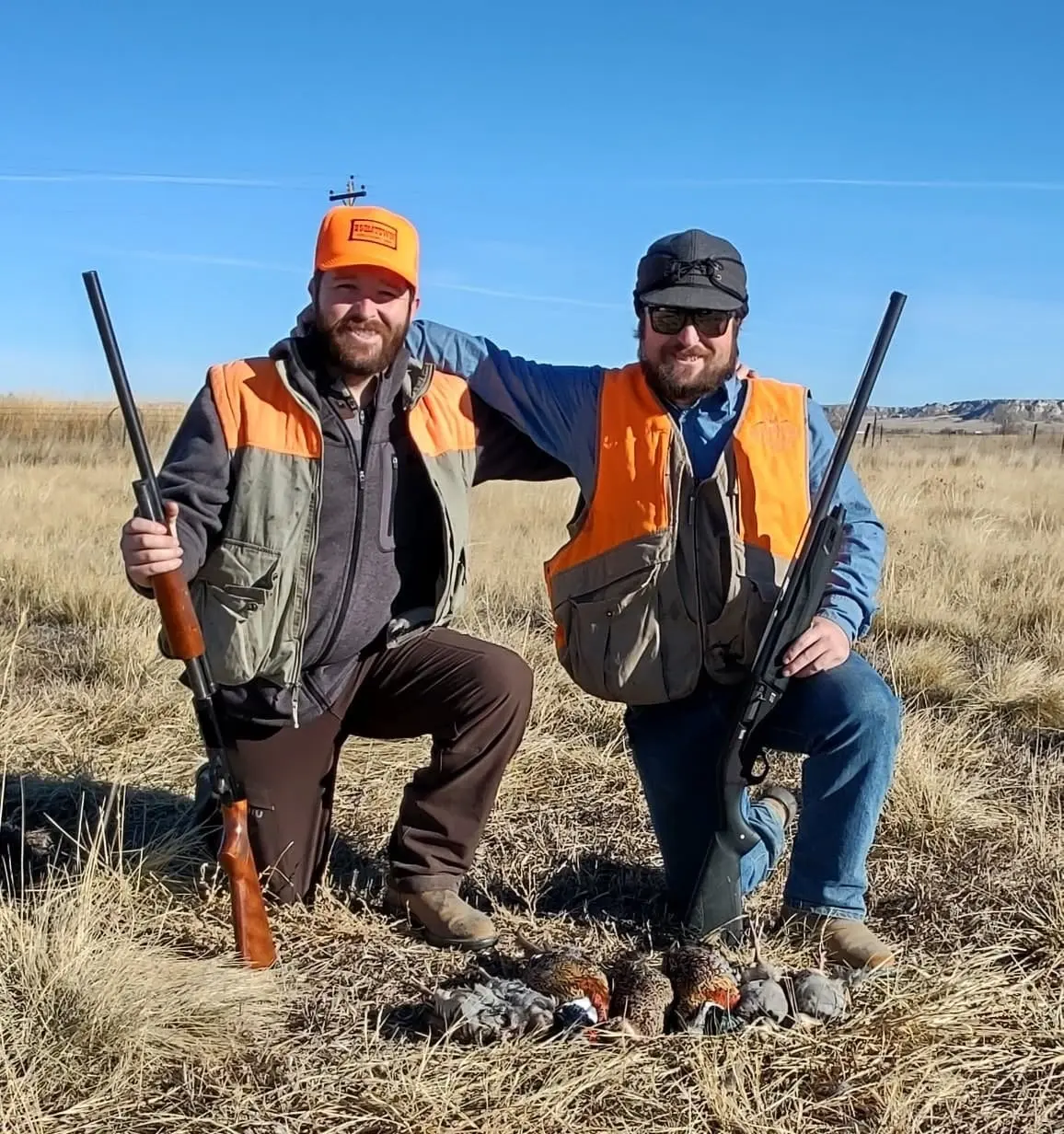 Two hunters pose with their shotguns and harvested pheasants and partridges in a field.