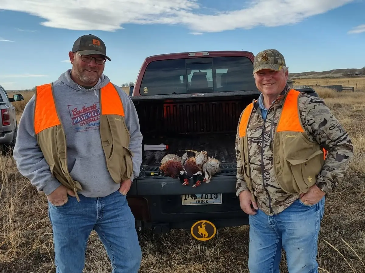 Two hunters pose with harvested pheasants and chukar in the back of a pickup truck.