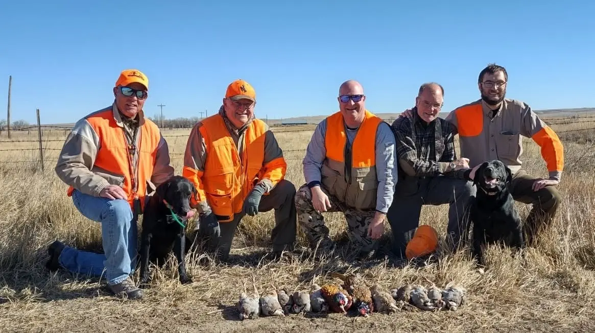 Five hunters and two dogs pose with their harvested pheasants and partridges in a field.