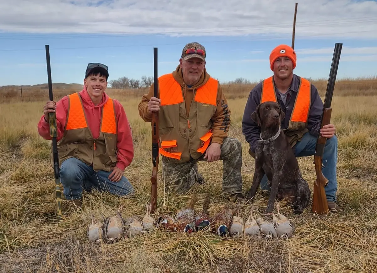 Three hunters and a dog pose with harvested Ring-necked Pheasants and Chukar Partridges in a field.