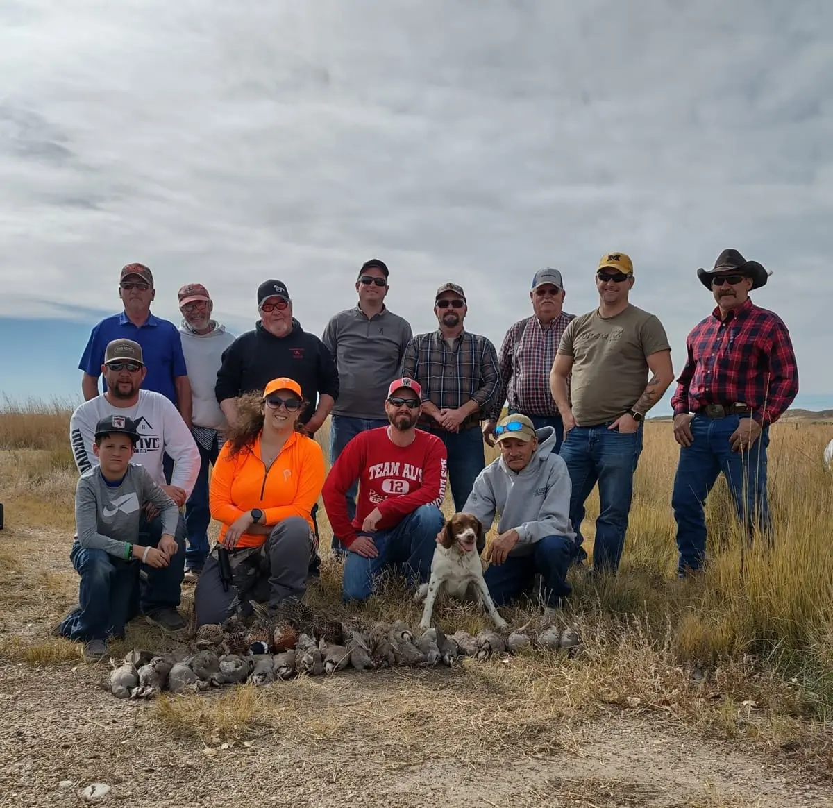 A group of hunters, a child, and a dog pose with a pile of harvested chukar partridges in a field.