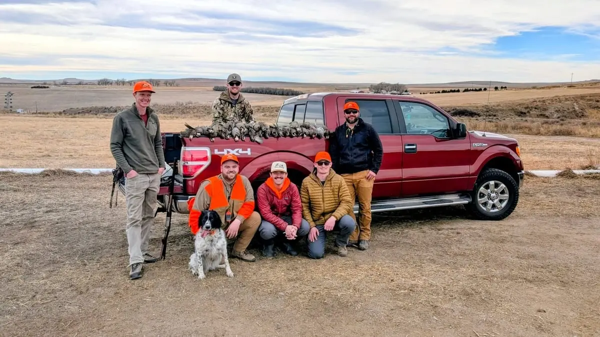 Several hunters and a dog pose with a truck full of harvested chukar in a field.