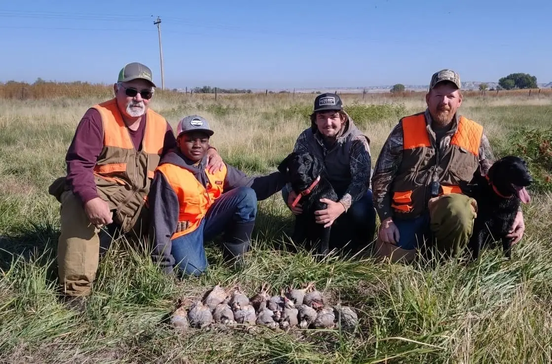Four hunters and two dogs pose with harvested chukar partridges in a grassy field.