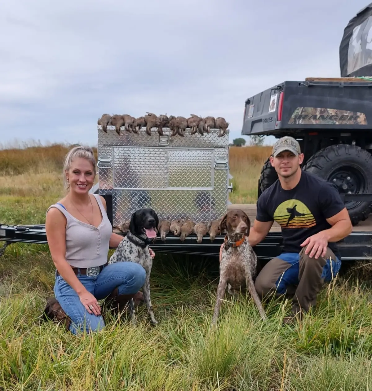 A man and a woman pose with two hunting dogs and a harvest of chukar next to a utility vehicle.