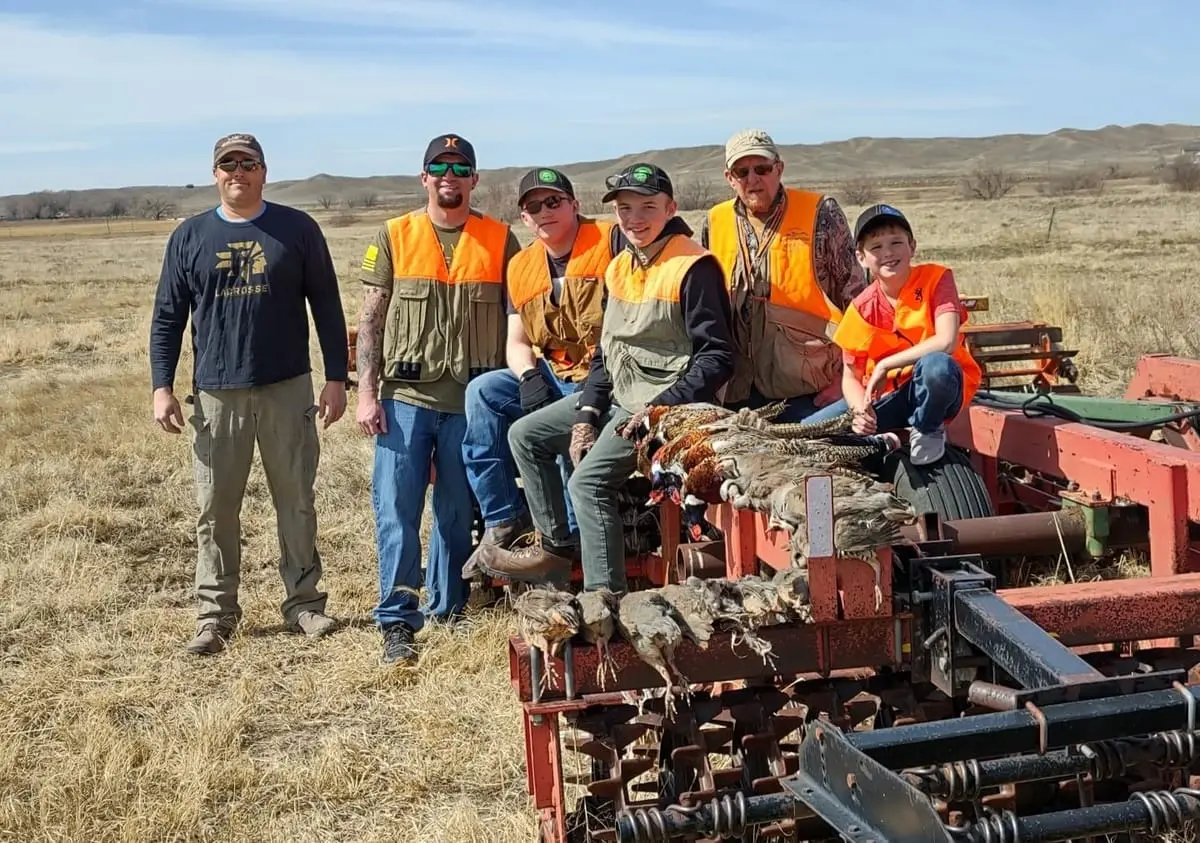 A group of hunters and a child pose with harvested pheasants and chukar on farm equipment in a field.