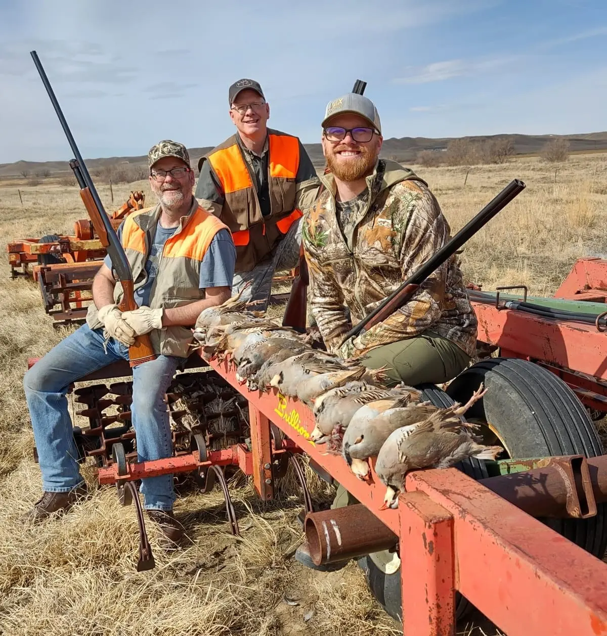 Three hunters pose with their harvested chukar partridges on a piece of farm equipment in a field.