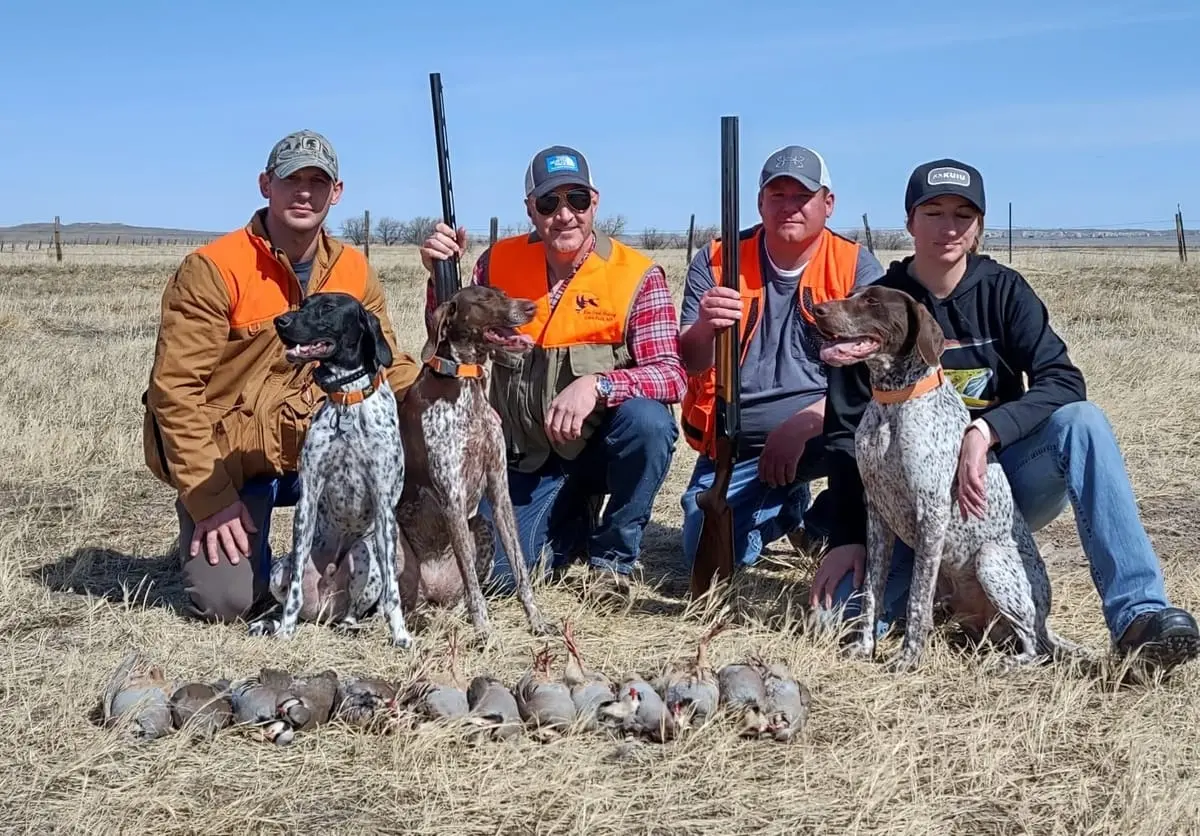 Four hunters and two dogs pose with harvested chukar partridges in a field.