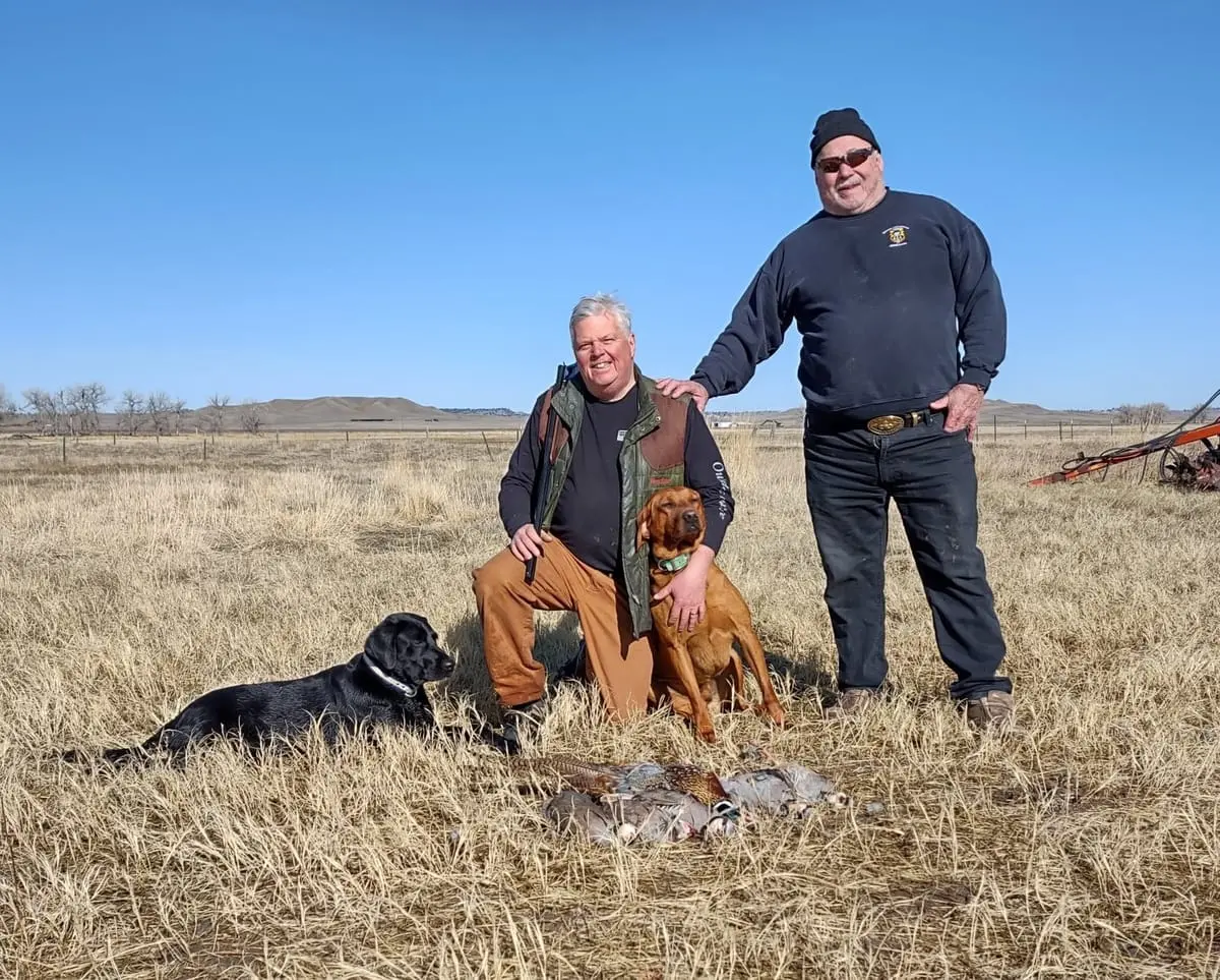 Two men and two dogs pose with harvested pheasants and chukar in a field.