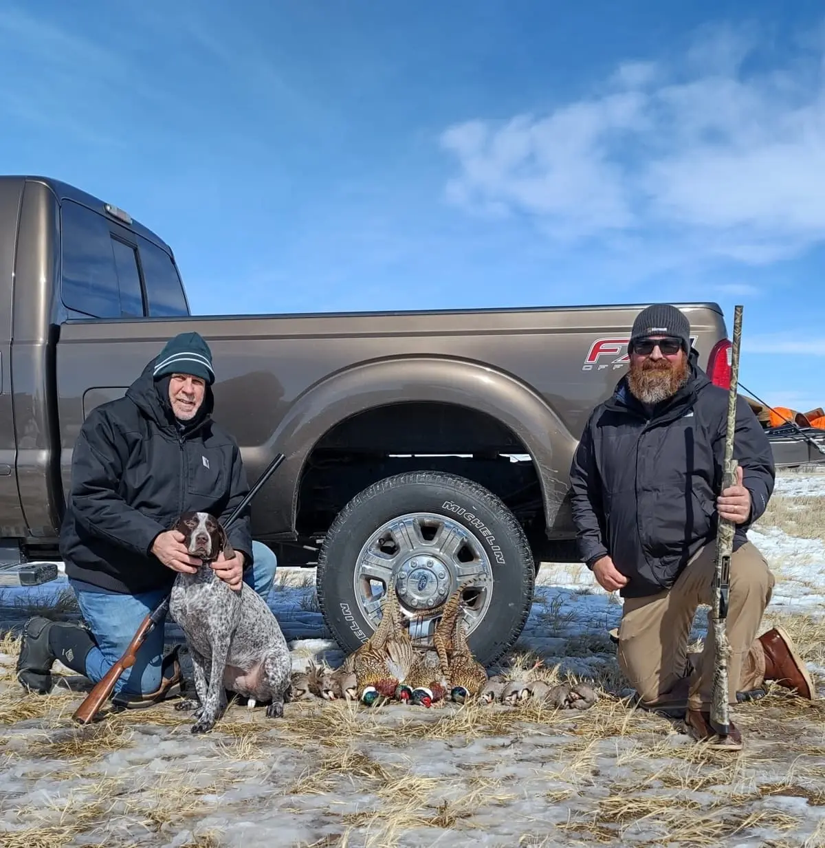 Two hunters and a dog pose with harvested pheasants and chukar next to a pickup truck.