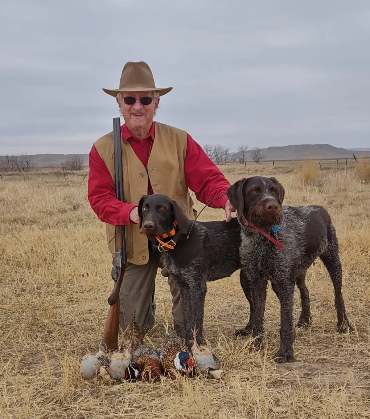 A hunter poses with two hunting dogs, harvested pheasants, and chukar partridges in a field.