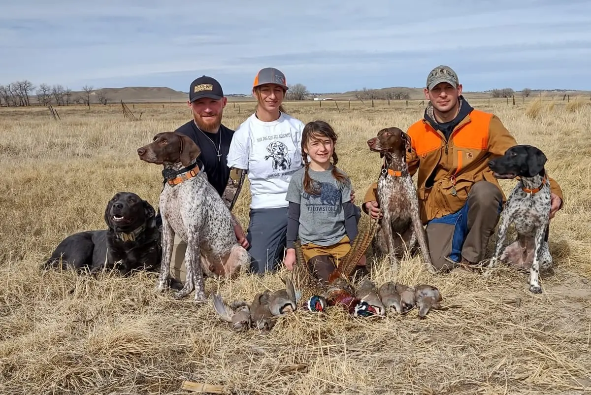 A group of hunters and a child, accompanied by four dogs, pose with harvested ring-necked pheasants and chukar partridges in a grassy field.