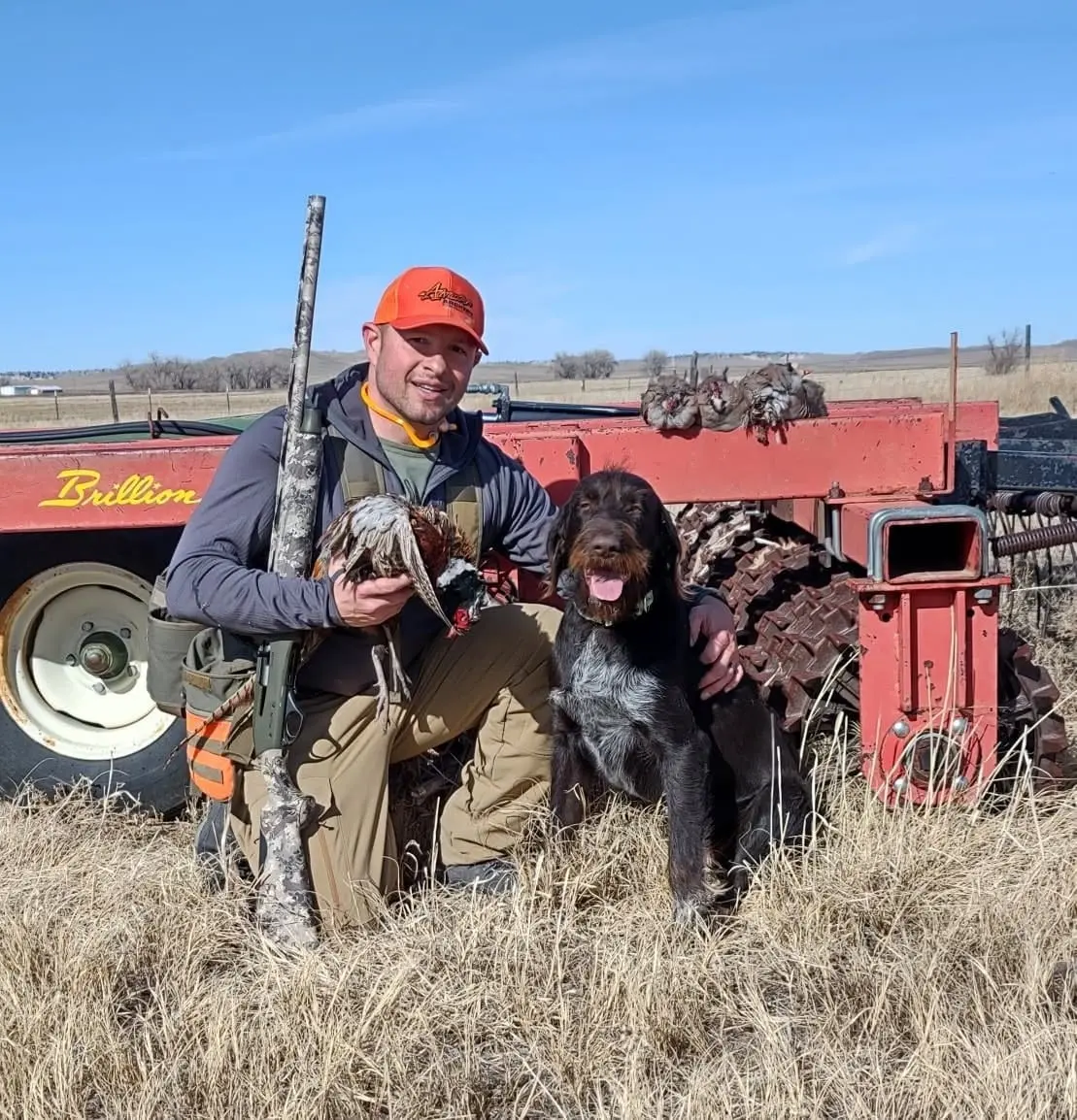 A hunter kneels with a harvested ring-necked pheasant and a hunting dog in a field.