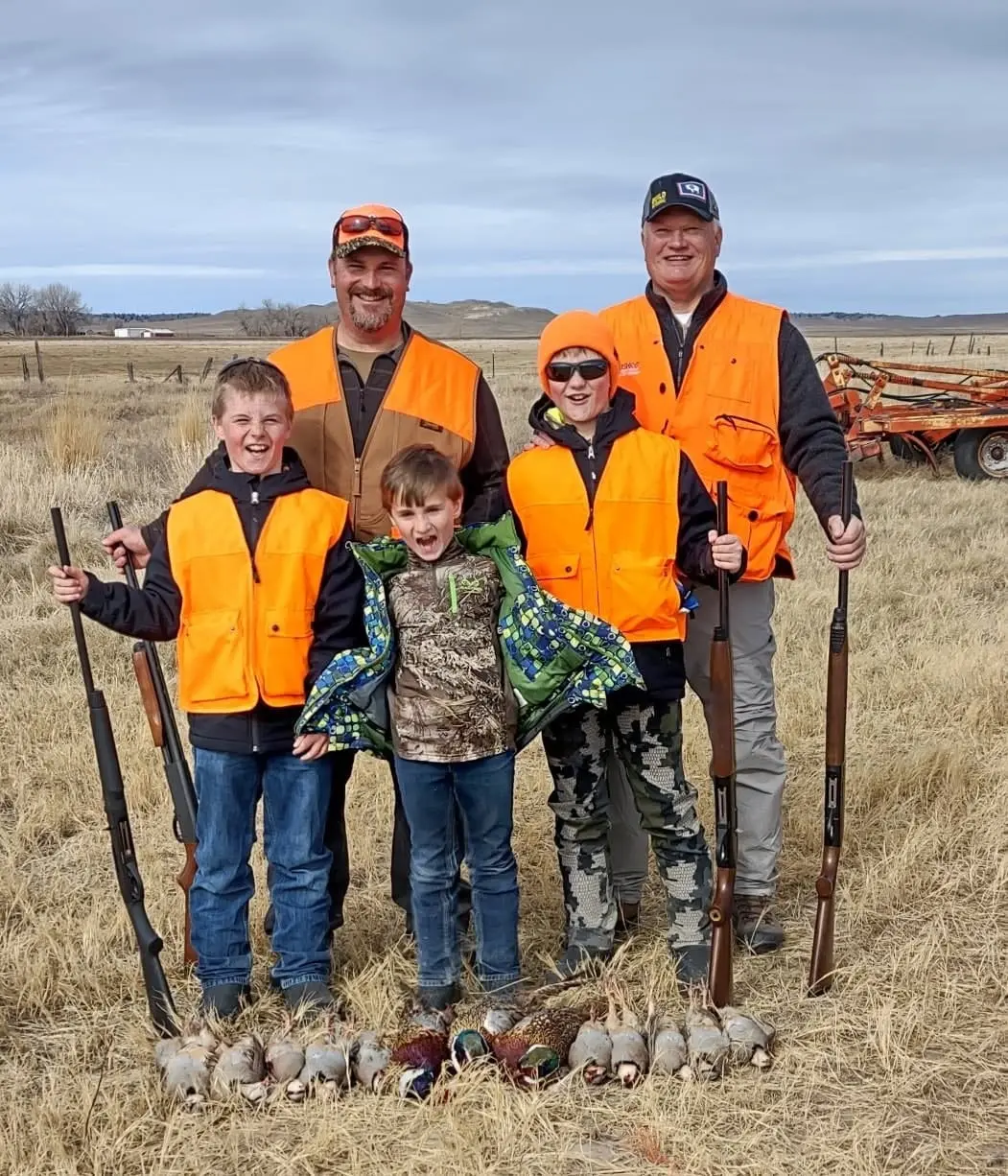 Several hunters, including children, pose with shotguns and harvested pheasants and chukar in a field.