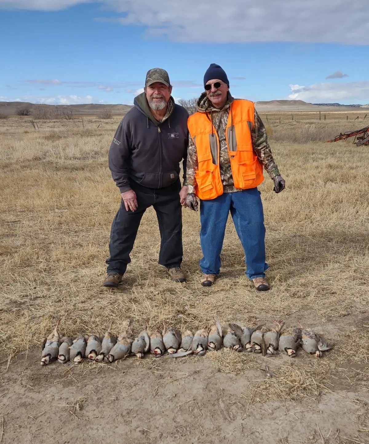 Two hunters pose with their harvested Chukar Partridges laid out on the ground in a field.