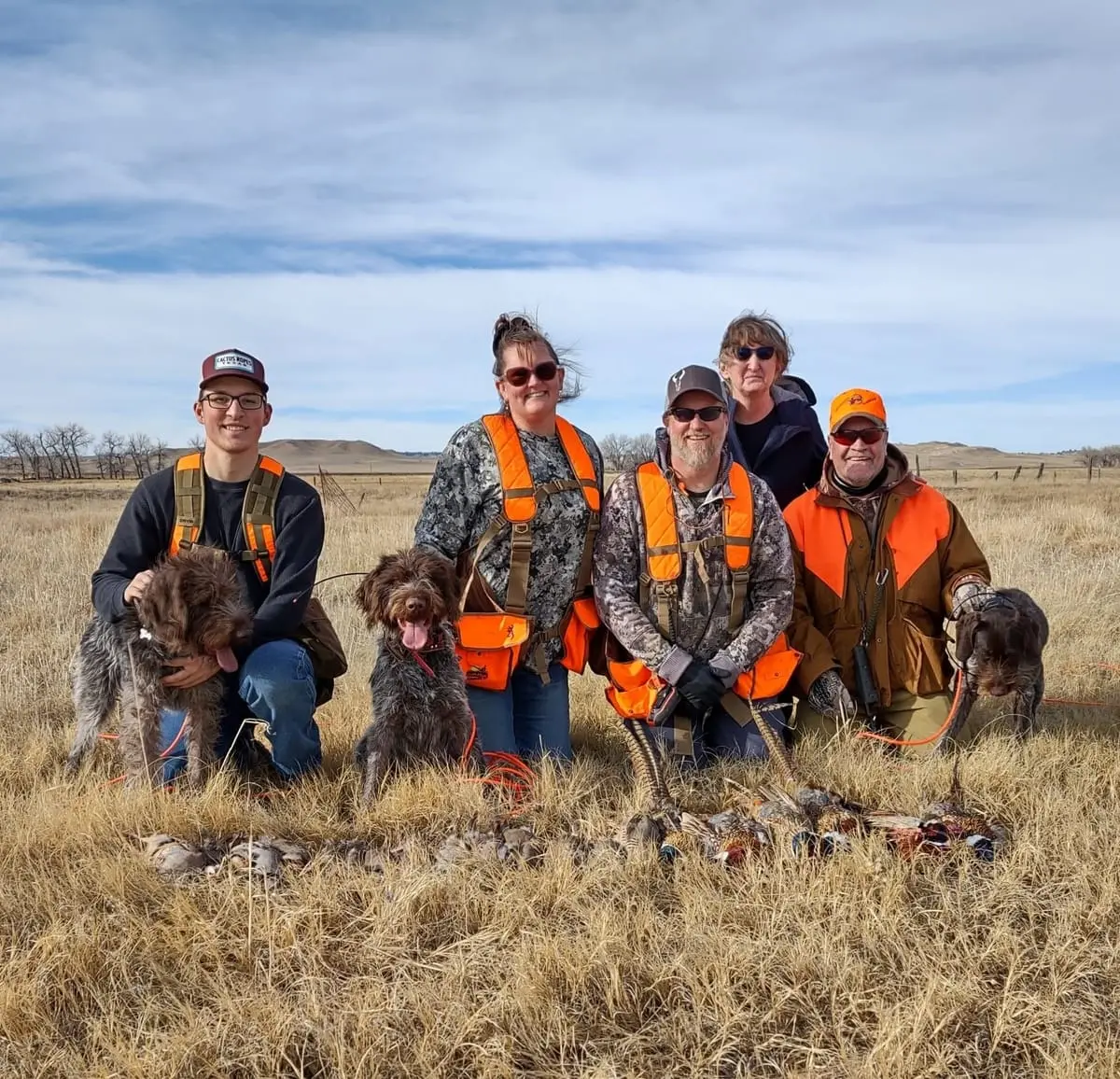 A group of hunters and their dogs pose with harvested pheasants and chukar in a grassy field.