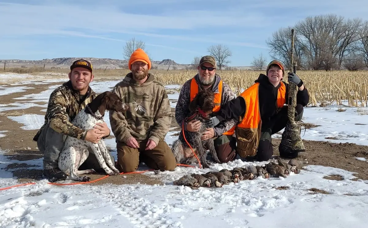 Four hunters and two dogs pose with their harvested chukar partridges in a snowy field.