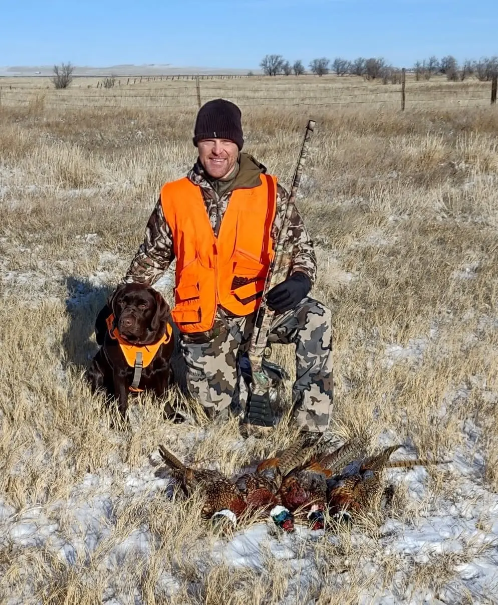A hunter and his dog pose with several harvested Ring-necked Pheasants in a snowy field.