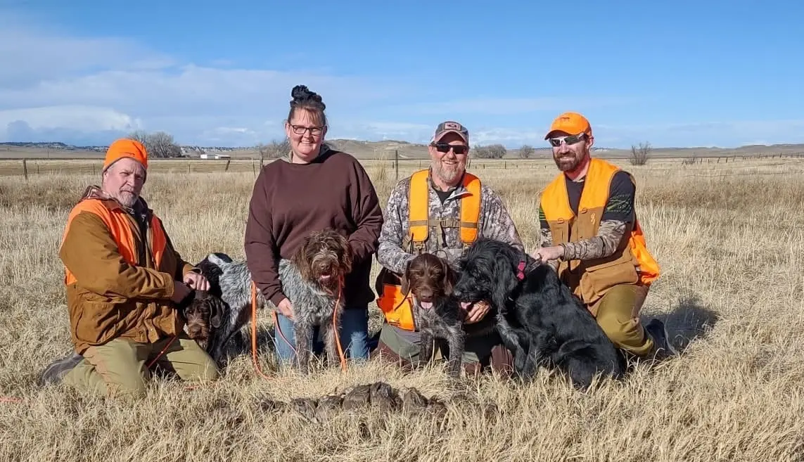Four hunters and five hunting dogs pose in a grassy field under a blue sky.