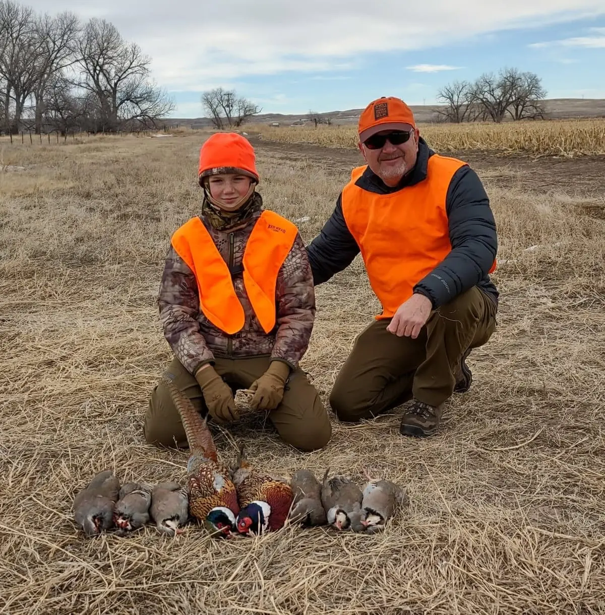 An adult and a child hunter pose with harvested ring-necked pheasants and chukar partridges in a field.