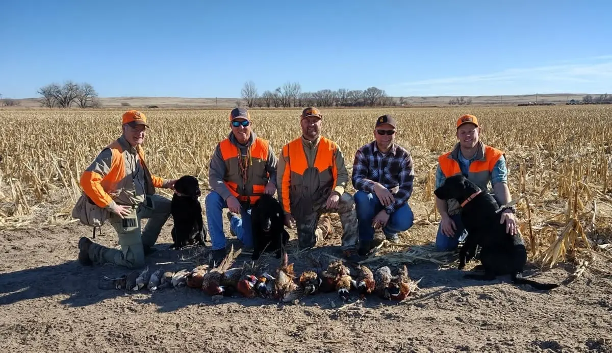 Five hunters and two dogs pose with harvested pheasants and chukar in a field.