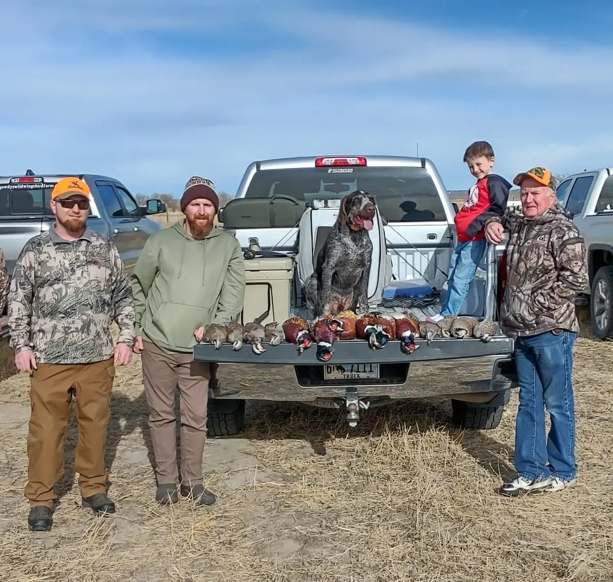A group of hunters, a child, and a dog pose with harvested pheasants and chukar in the back of a pickup truck.