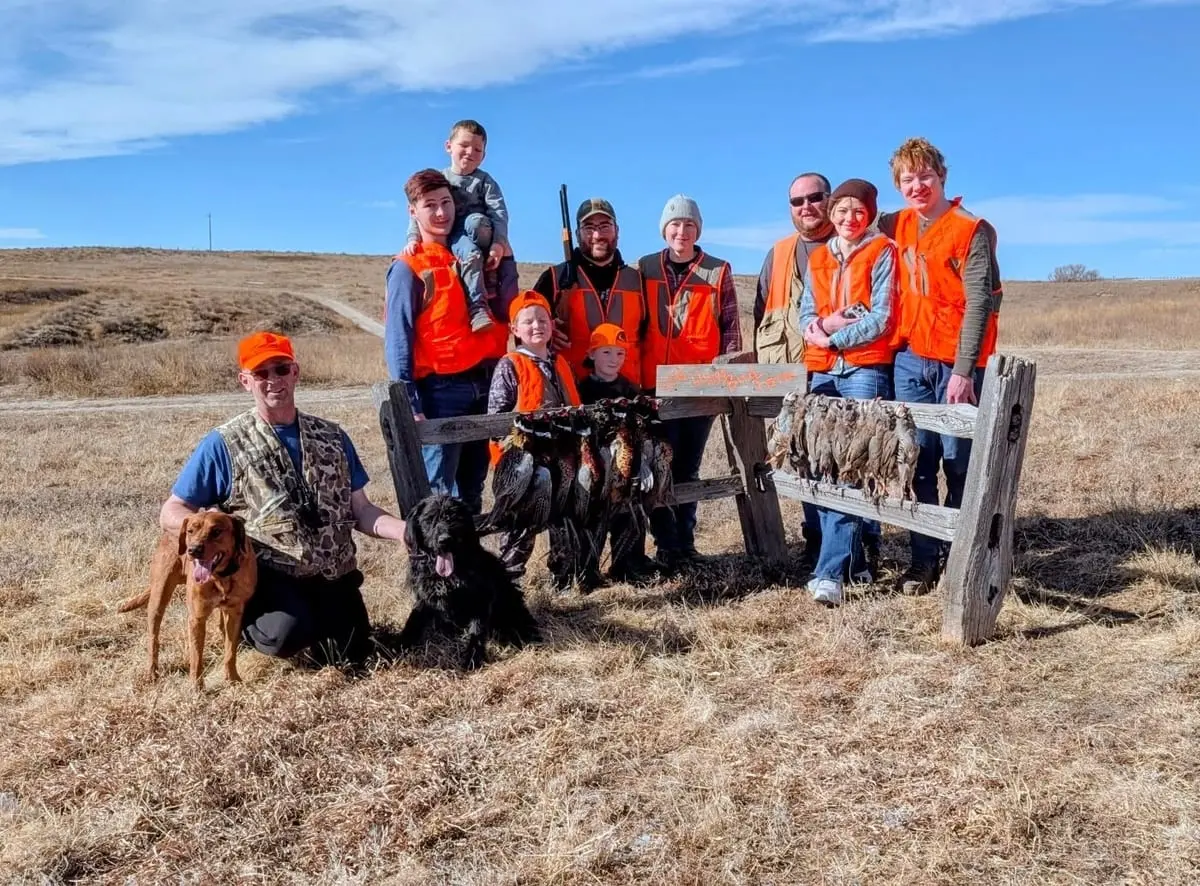 A group of hunters, including children, and their dogs pose with harvested pheasants and chukar on a sunny day.