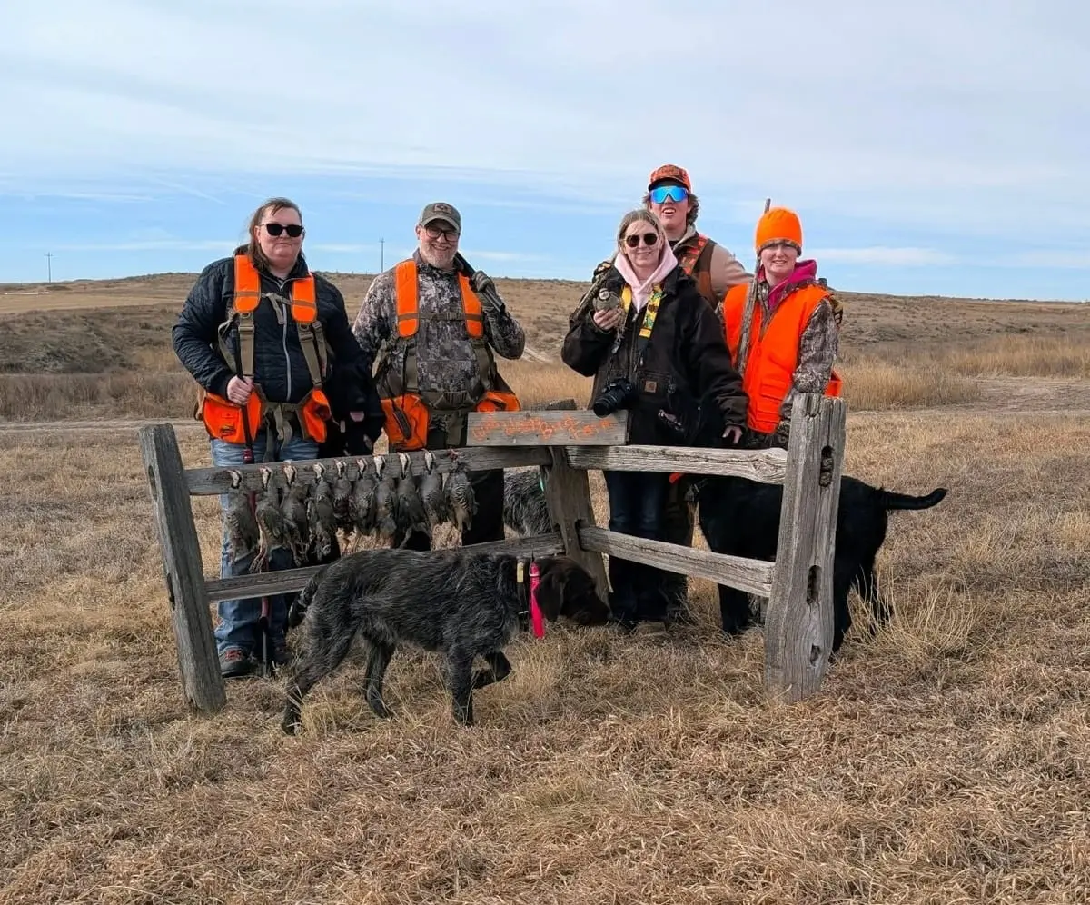 A group of hunters and two dogs pose with harvested chukar partridge hanging on a wooden rack in a field.