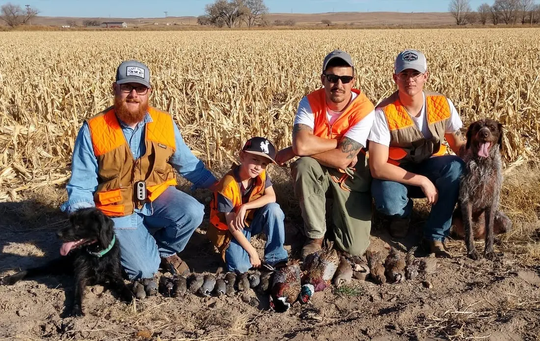 Three hunters, a boy, and two dogs pose with harvested pheasants and chukar in a field.