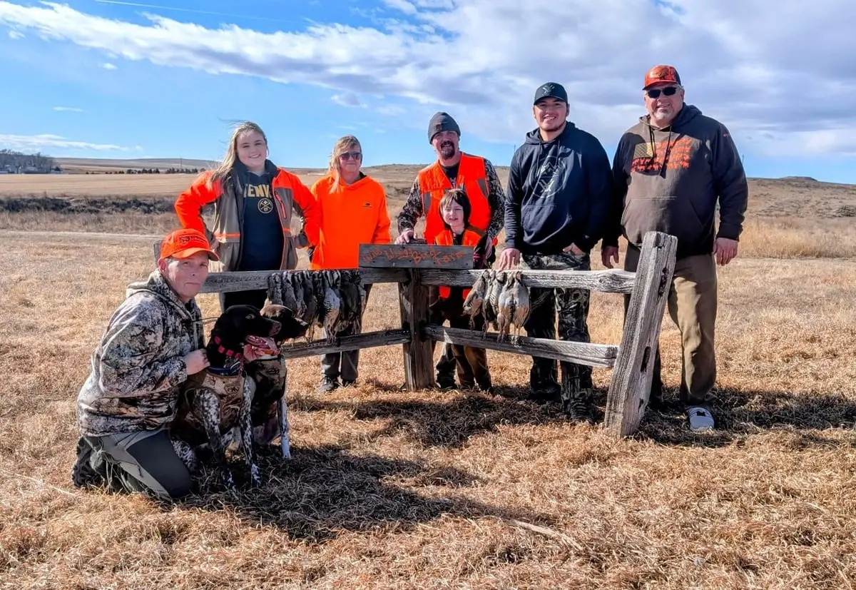A group of hunters, including a child, and a hunting dog pose with harvested chukar next to a wooden fence in a field.