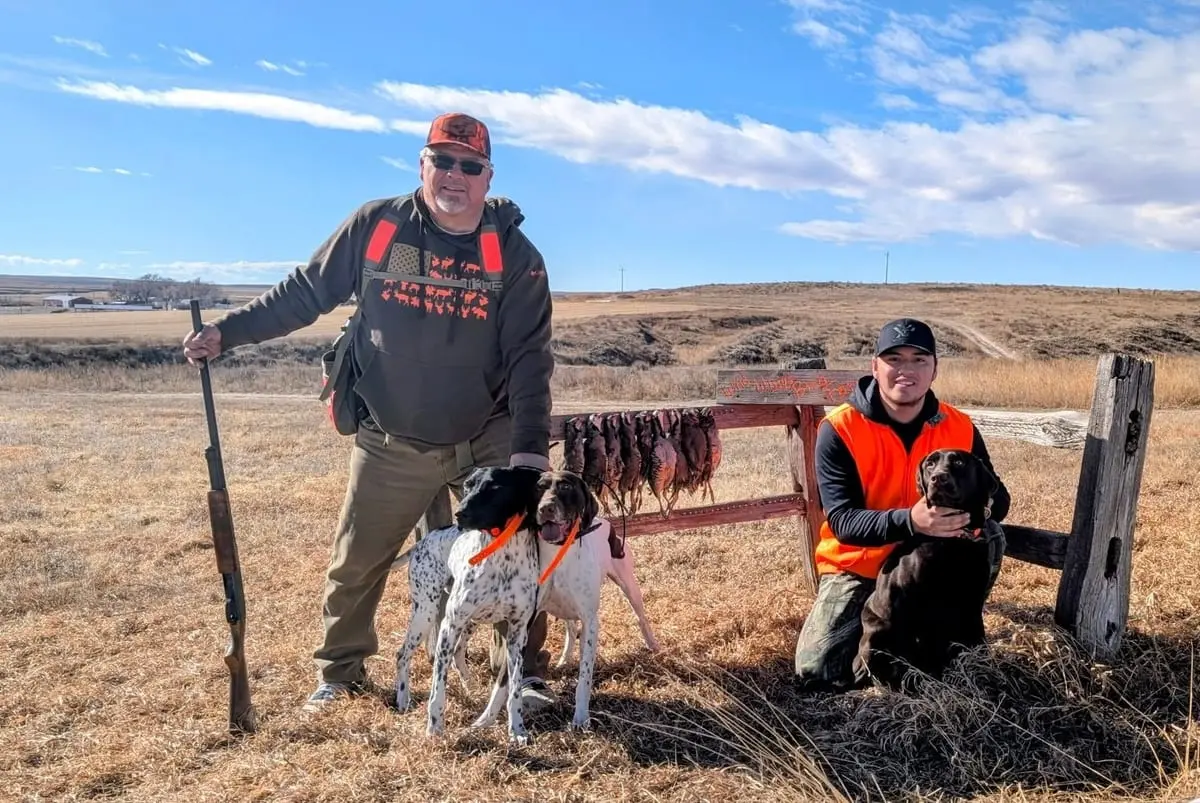 Two hunters pose with their dogs and harvested chukar hanging on a fence in a field.