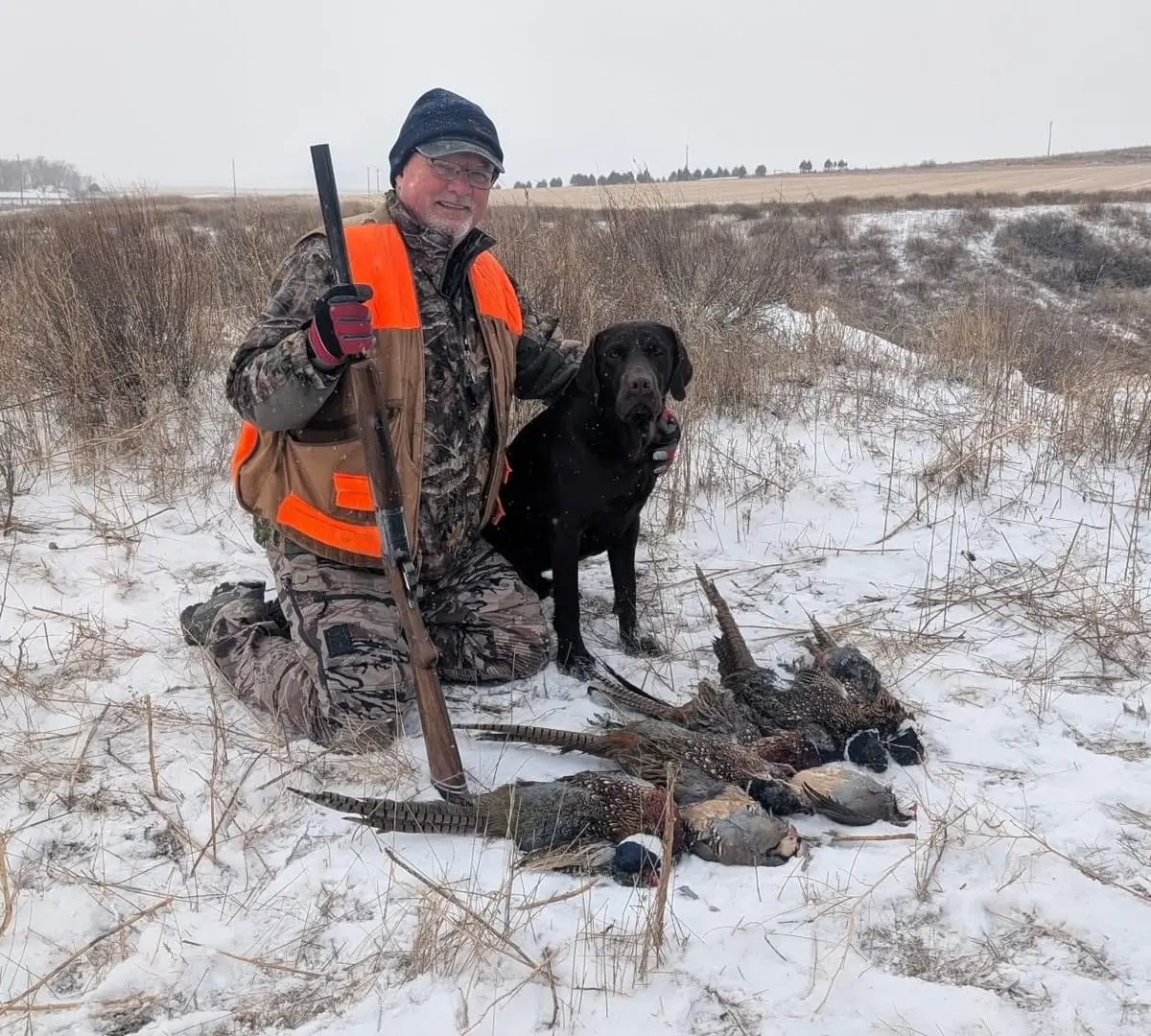 A hunter kneels in a snowy field with his black dog and several harvested pheasants and chukar.