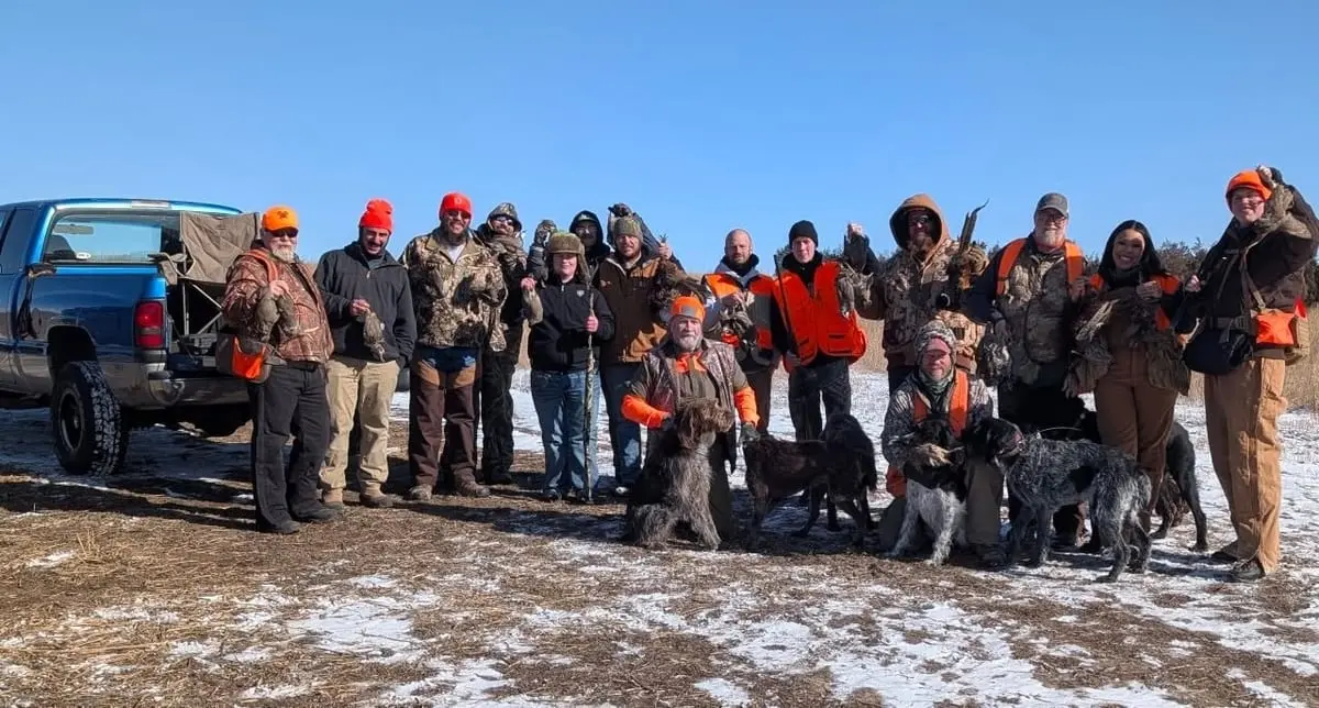 A group of hunters and their dogs pose with harvested ring-necked pheasants in a snowy field.