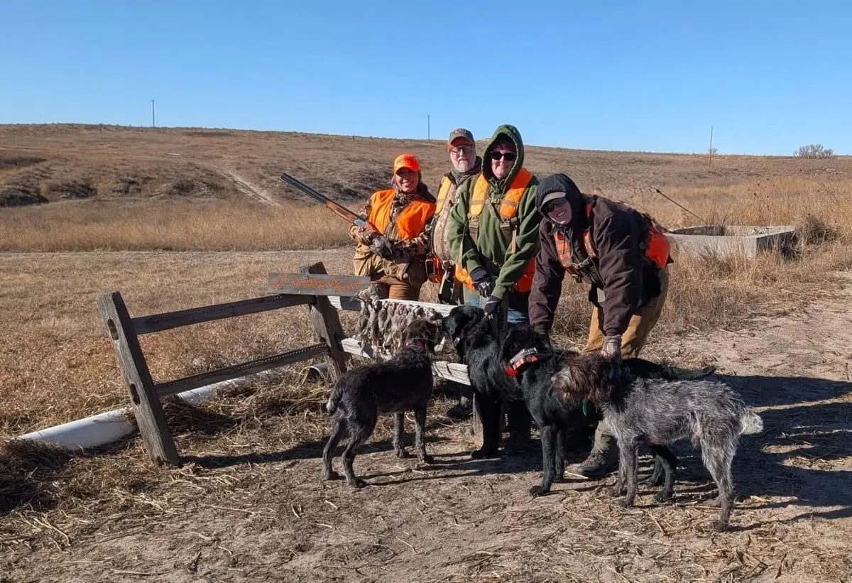 Hunters pose with harvested chukar and hunting dogs in a field.
