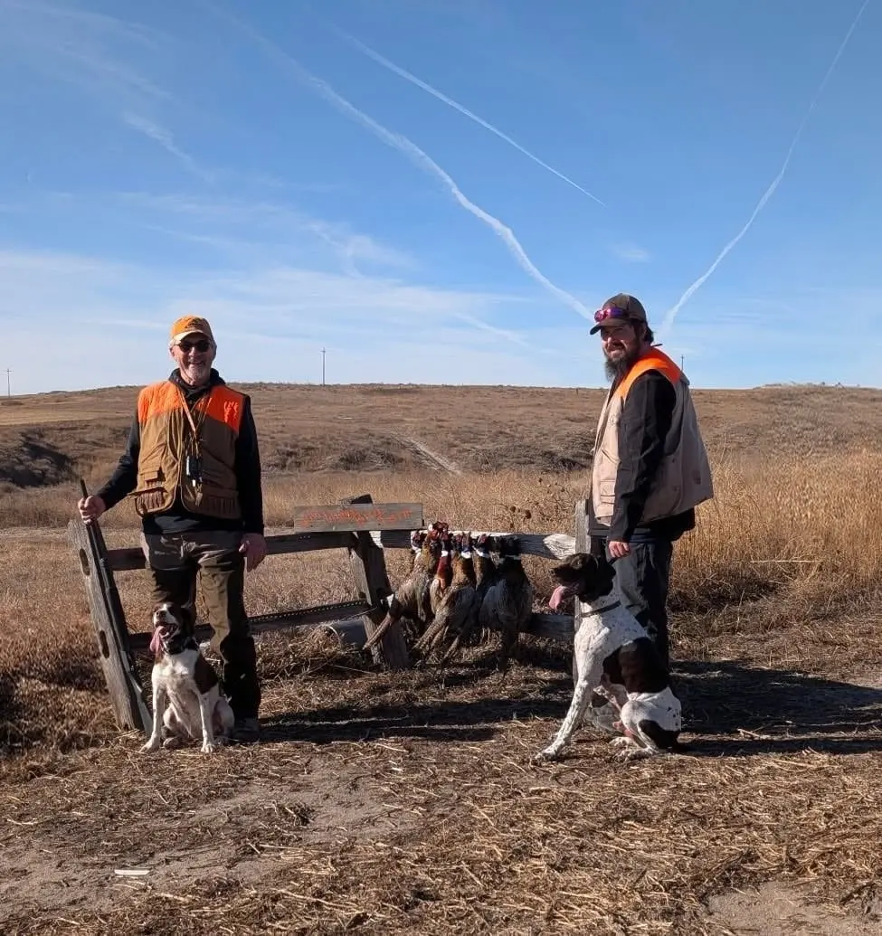 Two hunters pose with their dogs and harvested ring-necked pheasants in a field under a blue sky.