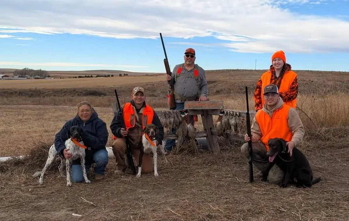 A group of hunters and their dogs pose with harvested pheasants and chukar in a field.