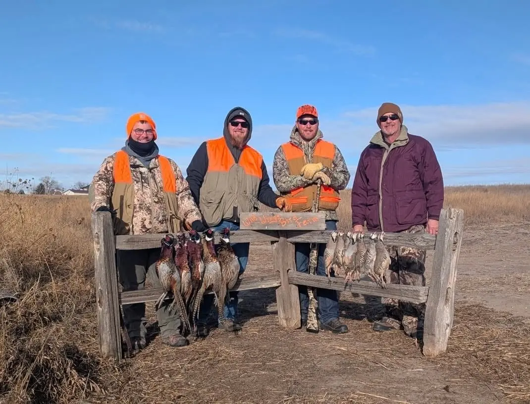 Four hunters pose with their harvested pheasants and chukar on a sunny day in a field.